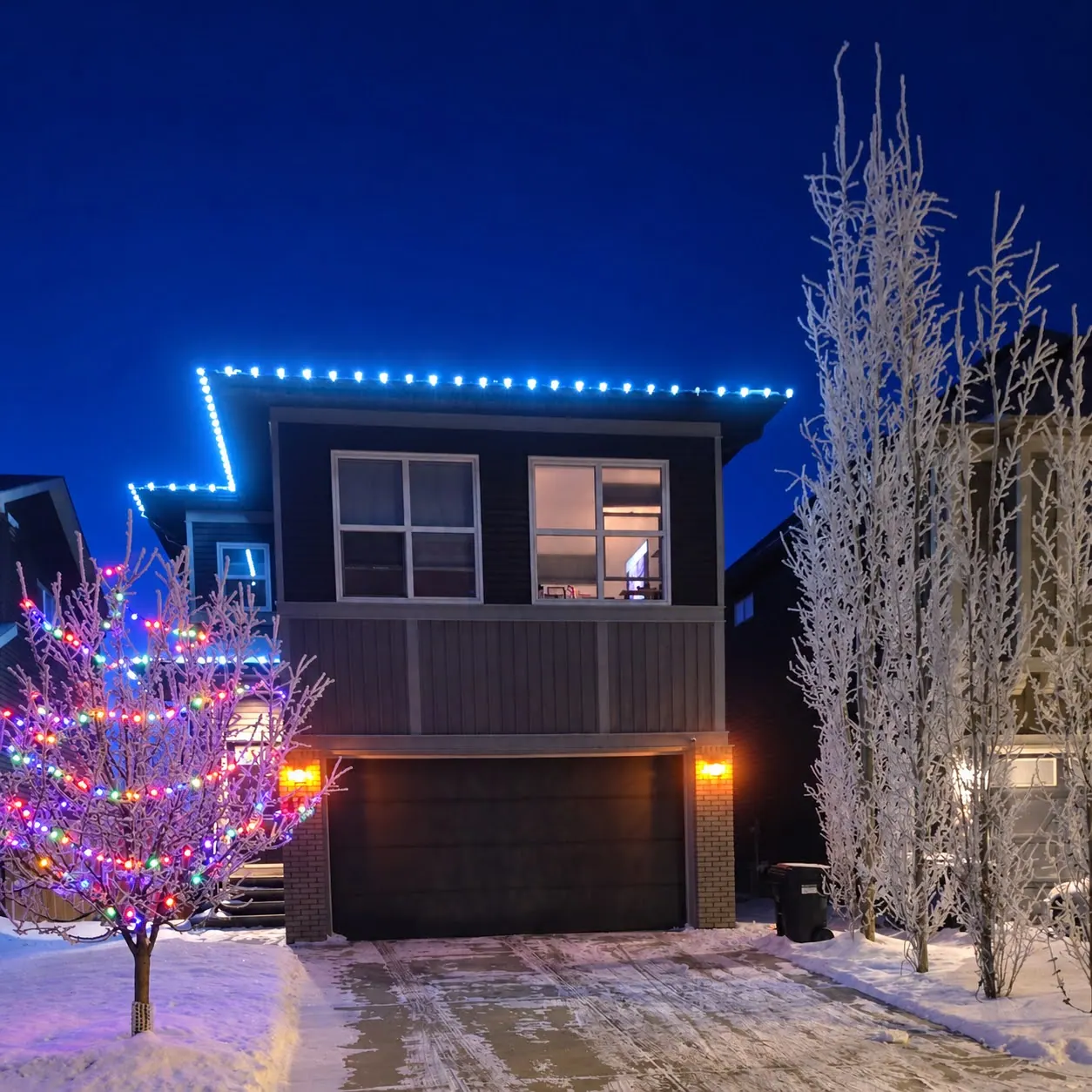 Modern two-storey home with ice blue bulbs along the roofline and a small tree wrapped in multicolour lights, at dusk